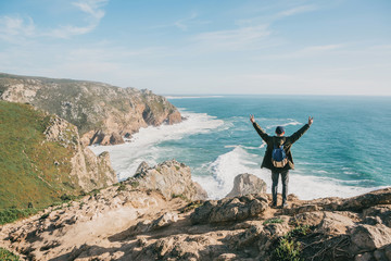 Wall Mural - A tourist with a backpack on a cliff against the background of the Atlantic Ocean raised his hands and shows how he is free and happy.