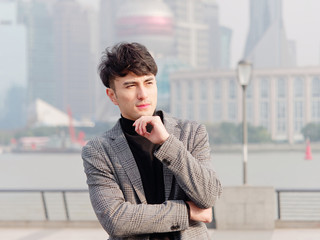 Portrait of a handsome Chinese young man in casual suit thinking with hand on chin with Shanghai bund background, winter fashion, cool young man lifestyle.