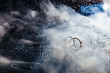 Wedding rings on dark surface shining with light close up macro. Water splashes. Reflection. Lights and smoke.