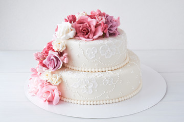 Two-tiered white wedding cake decorated with pink flowers on a white wooden background.