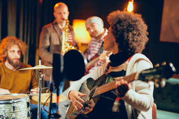 Sticker - Mixed race woman singing and playing guitar while sitting on chair.In background drummer, saxophonist and bass guitarist.