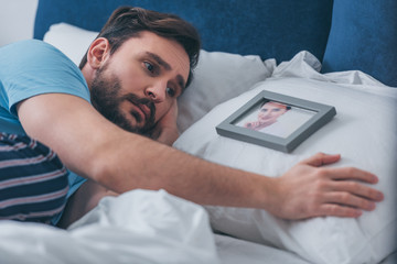 Wall Mural - man lying in bed and grieving near picture of woman on pillow at home