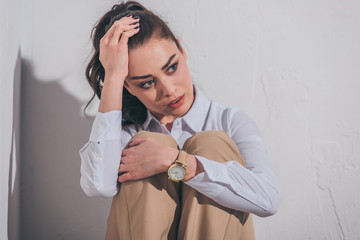 Wall Mural - sad woman in blouse and beige pants sitting on white background in room, grieving disorder concept