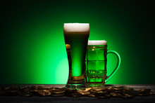 Glasses Of Irish Beer Standing Near Golden Coins On Wooden Table On Green Background