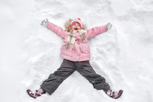 A Happy Little Toothless Girl In The Winter Walk, In The Nature. A Child Making An Angel On The Snow.