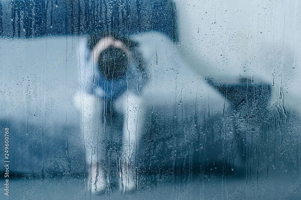 depressed woman sitting on bed and holding head in hands through window with raindrops - obrazy, fototapety, plakaty 