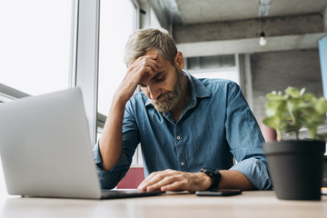 Wall Mural - Stylish modern man with a beard in a blue shirt is sitting at the table and working in a laptop in the office