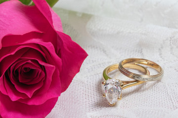 macro view of bright pink rose and narrow wedding band and diamond engagement ring on a white lace background