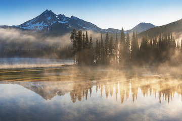 South Sister and Broken Top reflect over the calm waters of Sparks Lake at sunrise in the Cascades Range in Central Oregon, USA in an early morning light. Morning mist rises from lake into trees. 