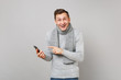 © ViDi Studio - Cheerful young man in gray sweater, scarf pointing index finger on mobile phone, typing sms message isolated on grey background. Healthy fashion lifestyle, people sincere emotions cold season concept.