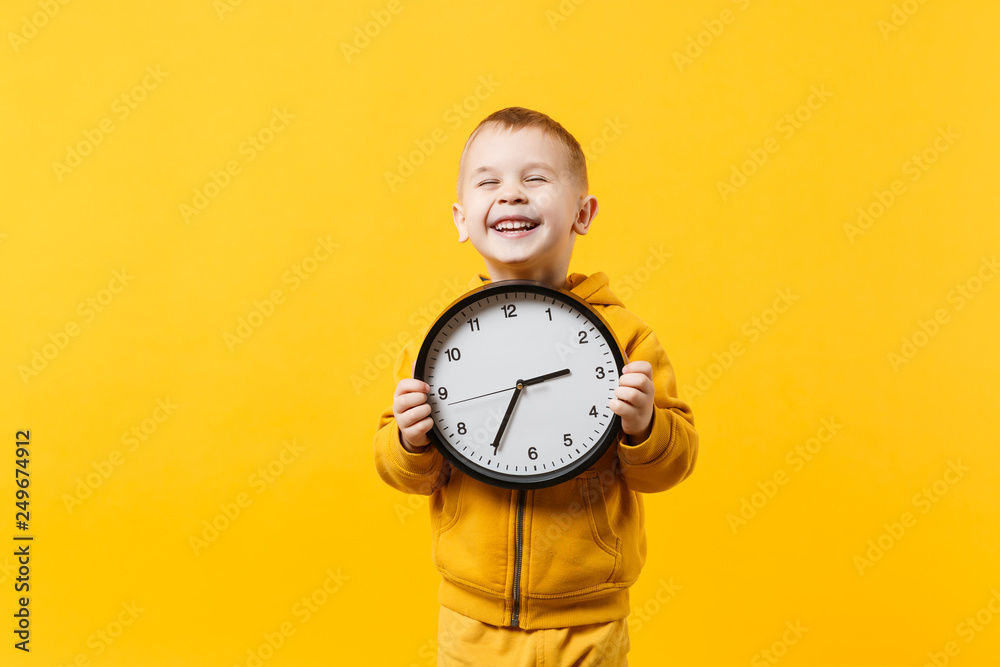 Little kid boy 3-4 years old wearing yellow clothes hold clock isolated on orange wall background, children studio portrait. People sincere emotions, childhood lifestyle concept. Mock up copy space. - obrazy, fototapety, plakaty 