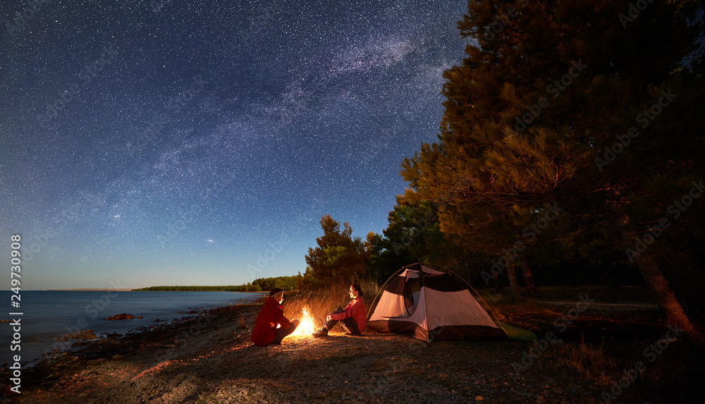 Night camping on shore. Young man and woman tourists sitting relaxed in front of tent at campfire under starry sky with Milky way on blue water and forest background. Active outdoor lifestyle concept - obrazy, fototapety, plakaty 