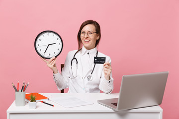 Wall Mural - Female doctor sit at desk work on computer with medical document hold money in hospital isolated on pastel pink wall background. Woman in medical gown glasses stethoscope. Healthcare medicine concept.