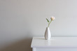 Close up of single pale pink rose in small vase on white sideboard against neutral wall background with copy space to left