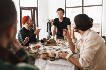 Young smiling asian man in eyeglasses and black T-shirt leaning on table happily looking in camera. Group of attractive international friends spending time together on lunch in cafe