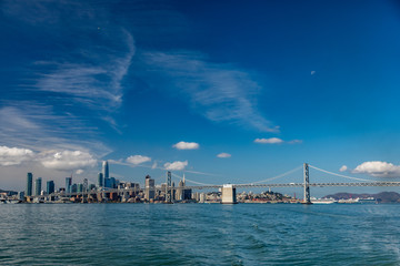 Wall Mural - Mostly blue skies fill the picture of the San Francisco skyline on the left and the bay bridge on the right with the moon in the upper right corner and water in the foreground