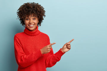 Cheerful young dark skinned lady smiles happily as shows pleasant thing, indicates with both fore fingers aside, dressed in loose red sweater, isolated over blue background. I like this one!