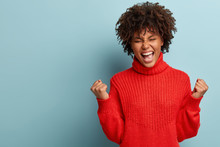 Success Is In My Pockets! Upbeat Overjoyed Woman Shows Victory Gesture, Raises Clenched Fists, Exclaims Happily, Cant Stop Positive Feelings, Wears Red Jumper, Isolated Over Blue Wall With Copy Space