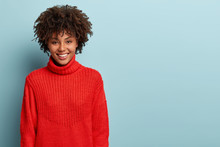 Sincere Feminine Girl With Dark Skin, Crisp Hair, Broad Smile, Has Fun, Enjoys Evening In Good Company, Wears Red Sweater With Collar, Models Over Blue Background With Free Space On Right Side