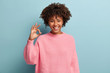 Headshot of positive dark skinned woman with crisp hair, makes okay gesture, wears loose sweater, enjoys life, isolated over blue background, says ok, confirms information. Its excellent result