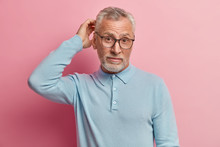 Shot Of Attractive Bearded Caucasian Man With Thick Grey Stubble, Scratches Head In Puzzlement, Wears Casual Blue Jumper And Spectacles, Isolated Over Rosy Background, Tries To Solve Problem