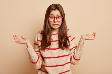 Wall Mural - Confused pretty girl has unaware facial expression, faces hard decision, wears glasses and jumper, feels unsure, clenches teeth, spreads hands sideways isolated over brown wall. Attitude concept