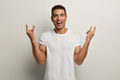 © WHstudio Leushin N - Happy pleased man with cheerful facial expression, makes rock n roll gesture, enjoys cool music, dressed in white t shirt, poses in studio, makes horns, has short dark hair. Body language concept