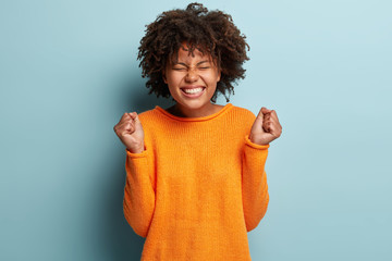Wall Mural - Oh yes finally I gained it! Overjoyed pleased dark skinned young woman raises clenched fists, shows white teeth, wears orange sweater, models over blue background, celebrates excellent news.
