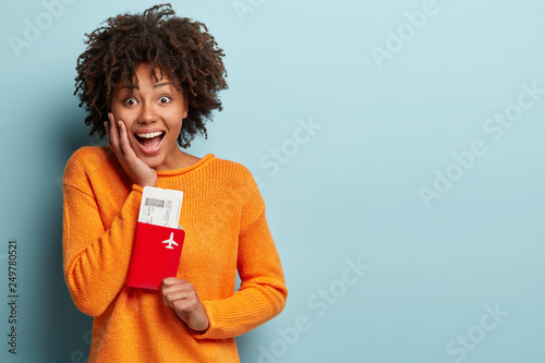 Happy pretty woman holds passport and plane tickets, rejoices having tour abroad with boyfriend, has special offer from travel company, isolated over blue background, wears knitted orange jumper