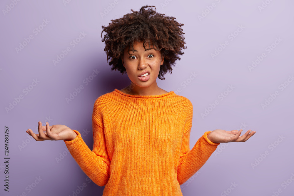 Isolated shot of confused beautiful woman with dark skin, curly haircut, spreads hands sideways, smirks face, feels doubt while makes choice, dressed in casual jumper, isolated over purple wall - obrazy, fototapety, plakaty 