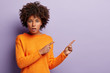 Shot of puzzled dark skinned girl in casual jumper, points with both fore fingers at empty space, shows something with indignation, isolated over lilac background. Scared lady advertises scary thing