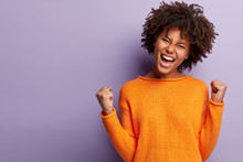 Life Is Wonderful! Joyful Dark Skinned Lady Keeps Fists Clenched, Tilts Head And Exclaims In Triumph, Celebrates Success, Isolated Over Purple Background With Blank Space For Your Advertisement