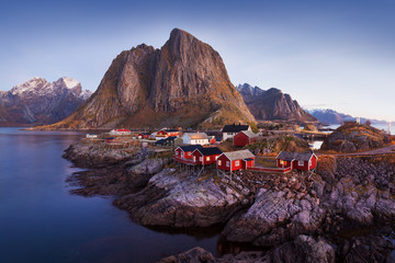 Wall Mural - Winter landscape with houses in village, city illumination, snowy mountains, sea, blue cloudy sky reflected in water at dusk. Beautiful Hamnoy, Lofoten islands, Norway. Norwegian fishing village