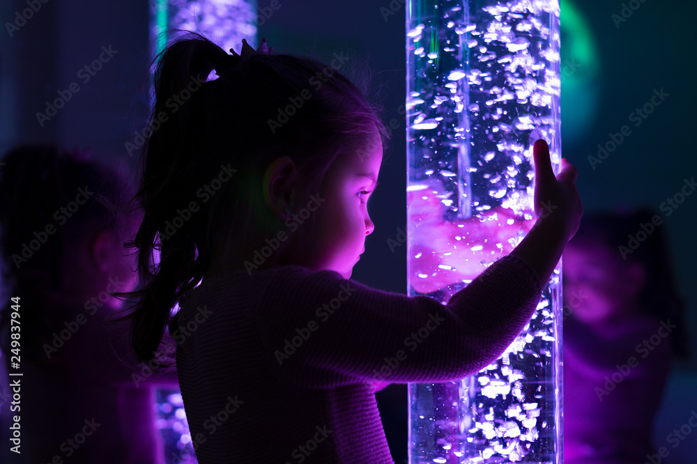 Child in therapy sensory stimulating room, snoezelen. Child interacting with colored lights bubble tube lamp during therapy session. - obrazy, fototapety, plakaty 