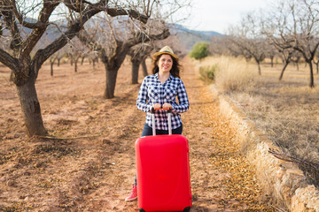 Wall Mural - Travel, tourism and people concept - happy young woman going to travel by with red suitcase and smiling over the nature