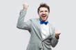 © khosrork - celebrating victory. Portrait of excited happy handsome bearded man in casual grey suit and blue bow tie standing with raised arms and satisfied. indoor studio shot, isolated on light grey background.