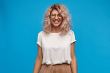 Emotional Charming Young European Female In Trendy Glasses Laughing, Closing Eyes And Smiling Broadly, Showing Her White Perfect Teeth. Attractive Girl In Good Mood Having Fun, Posing At Blue Wall