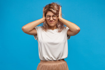 Wall Mural - Irritated angry female hipster in trendy eyewear covering ears with hands and screwing eyes being annoyed with squeaky sound or loud music. Frustrated teen girl can't stand her parents fighting