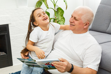 Wall Mural - Happy little girl with grandfather reading story book at home