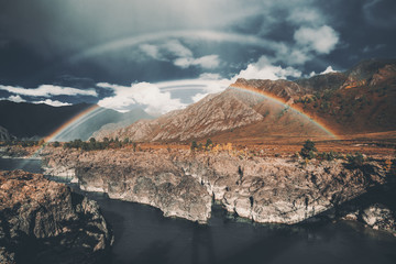 A stunning double rainbow over two banks of a mountain river Katun in Altai mountains, Russia: dark-blue stormy sky, rare clouds, autumn meadows overgrown with native grasses, cliffy riverside