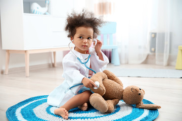 Wall Mural - Cute African American child imagining herself as doctor while playing with stethoscope and toy bunny at home