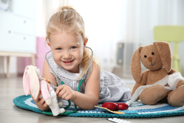 Wall Mural - Cute child imagining herself as doctor while playing with stethoscope and toy bunny at home