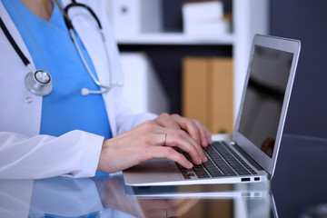 Doctor typing on laptop computer while sitting at the glass desk in hospital office. Physician at work. Medicine and healthcare concept