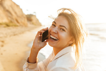 Sticker - Image of european girl 20s smiling and talking on mobile phone, while walking by seaside