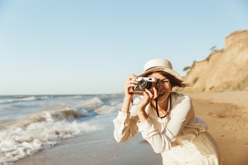 Poster - Image of cheerful woman 20s photographing on retro camera, while walking by seaside