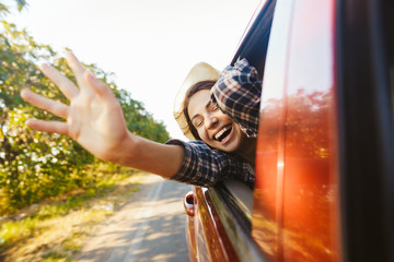 Wall Mural - Image of caucasian woman 20s wearing straw hat laughing and waving hand out of the window, while riding in car