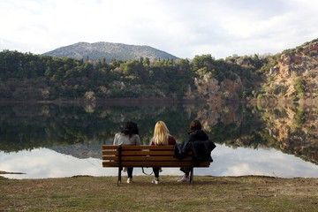 Three female young friends sitting on a wooden bench at lake shore staring at the view of lake Ziros with calm water the reflection and the forest at the background in Preveza Epirus 