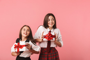 Portrait of lovely girls in school uniform holding present boxes, while standing isolated over red background