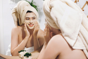 Young happy woman in towel making facial massage with  organic face scrub and looking at mirror in stylish bathroom. Girl applying scrub cream, peeling and cleaning skin. Skin Care