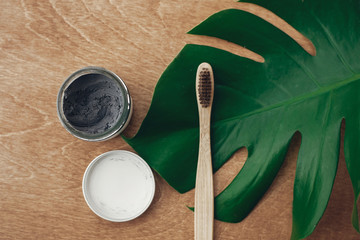 Natural toothpaste activated charcoal and bamboo toothbrush on wooden background with green monstera leaf. Plastic free beauty essentials, teeth care. Zero waste concept. Sustainable lifestyle.